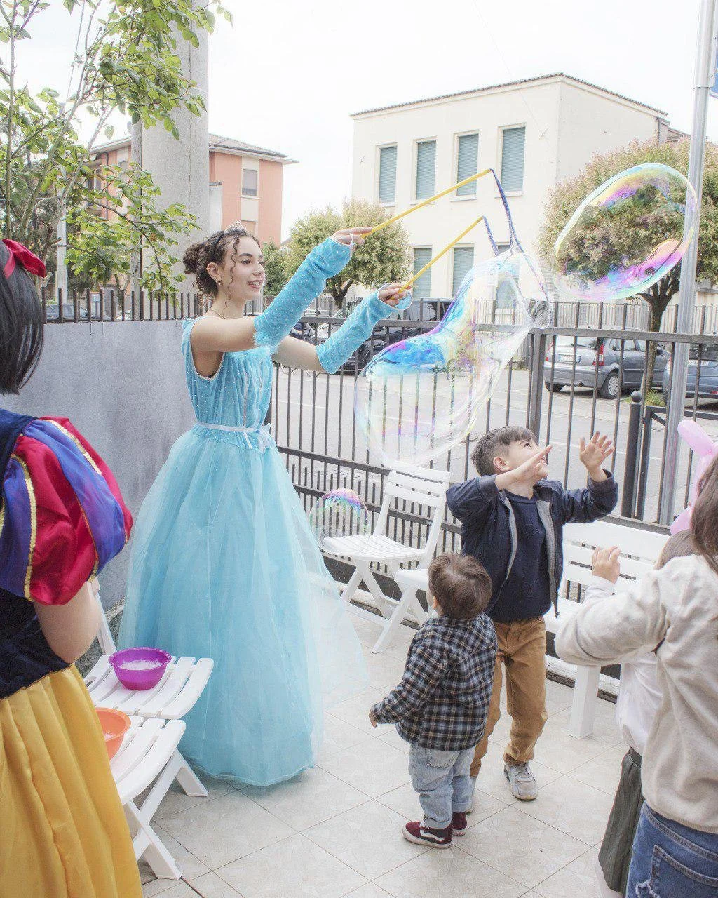 Una donna travestita da principessa crea grandi bolle di sapone con un bastoncino, mentre alcuni bambini la osservano e cercano di toccarle. L'evento si svolge all'aperto, in un cortile con sedie e una recinzione.
