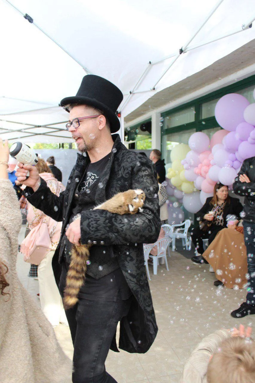 Uomo con cappello alto, occhiali e giacca nera con motivi floreali, che tiene un procione tra le braccia, durante una festa con palloncini color pastello e persone sedute e in piedi.