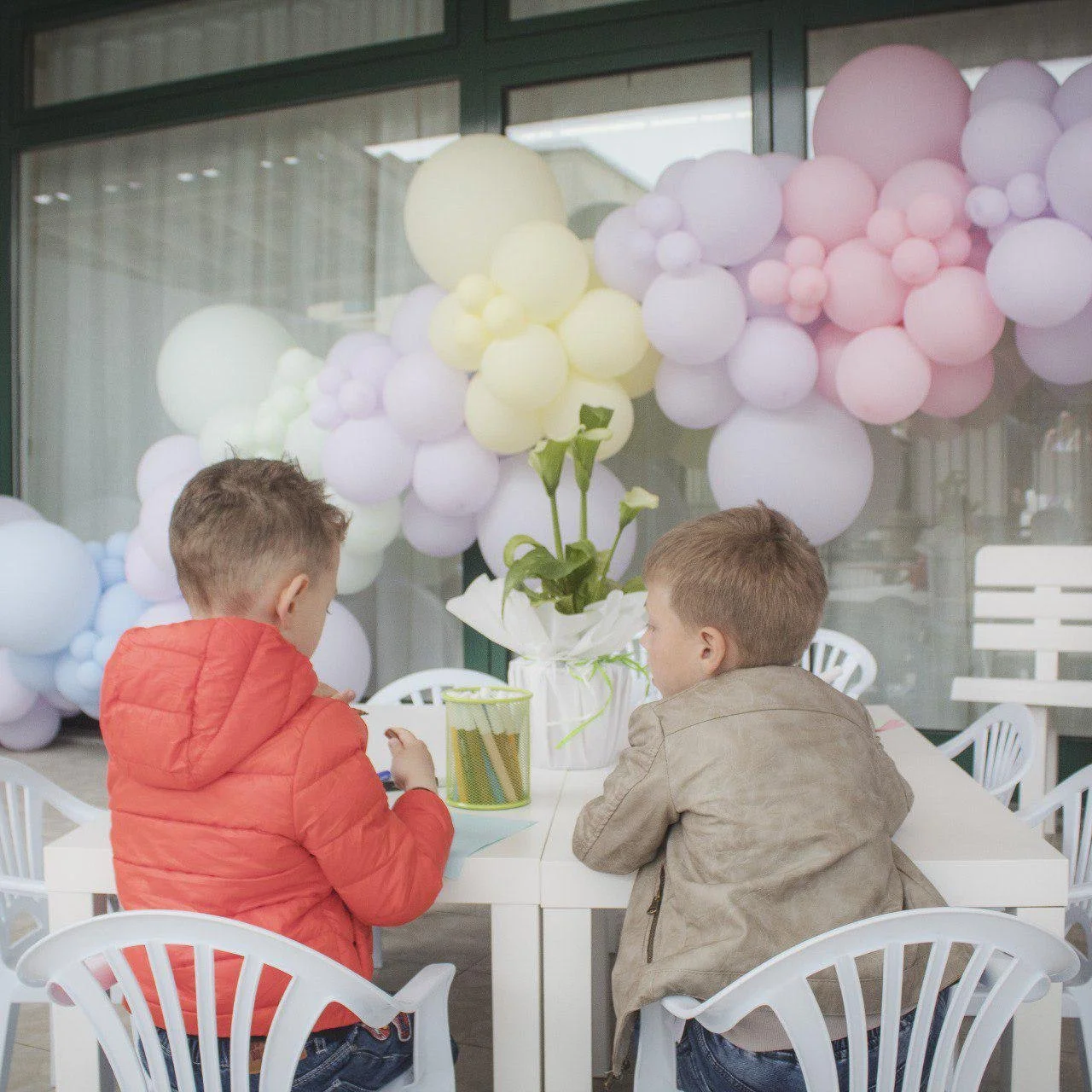 Due bambini seduti a un tavolo decorato con palloncini pastello, sembra una festa, uno indossa una giacca rossa e l'altro una beige.