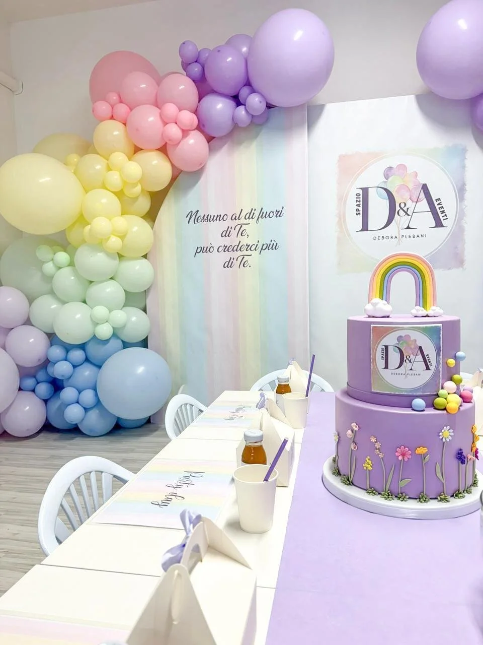 Decorazioni di compleanno con palloncini di diversi colori, una torta viola decorata con fiori e arcobaleno, e un muro con scritta in italiano. Ci sono anche tovagliette colorate e bottiglie di bibite sulla tavola.