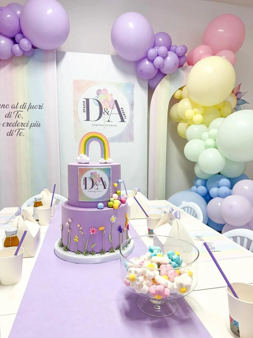 Torta decorata con motivi floreali e arcobaleno pastello, circondata da palloncini colorati e decorazioni per festa. Tavola con ciotola di caramelle colorate e lattine di bibite.