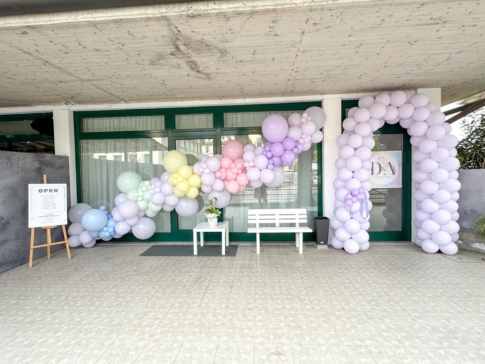 Decorazione con palloncini color pastello in un'entrata di locale, con porta in vetro, panchina bianca, vaso con fiori e cartello 'Open' vicino.