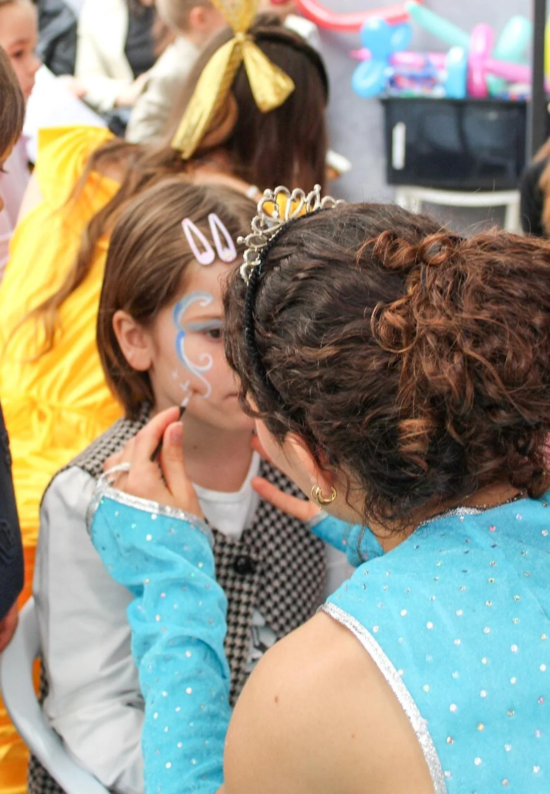 Una donna sta facendo il trucco a un bambino in un evento, con altri bambini e adulti sullo sfondo.