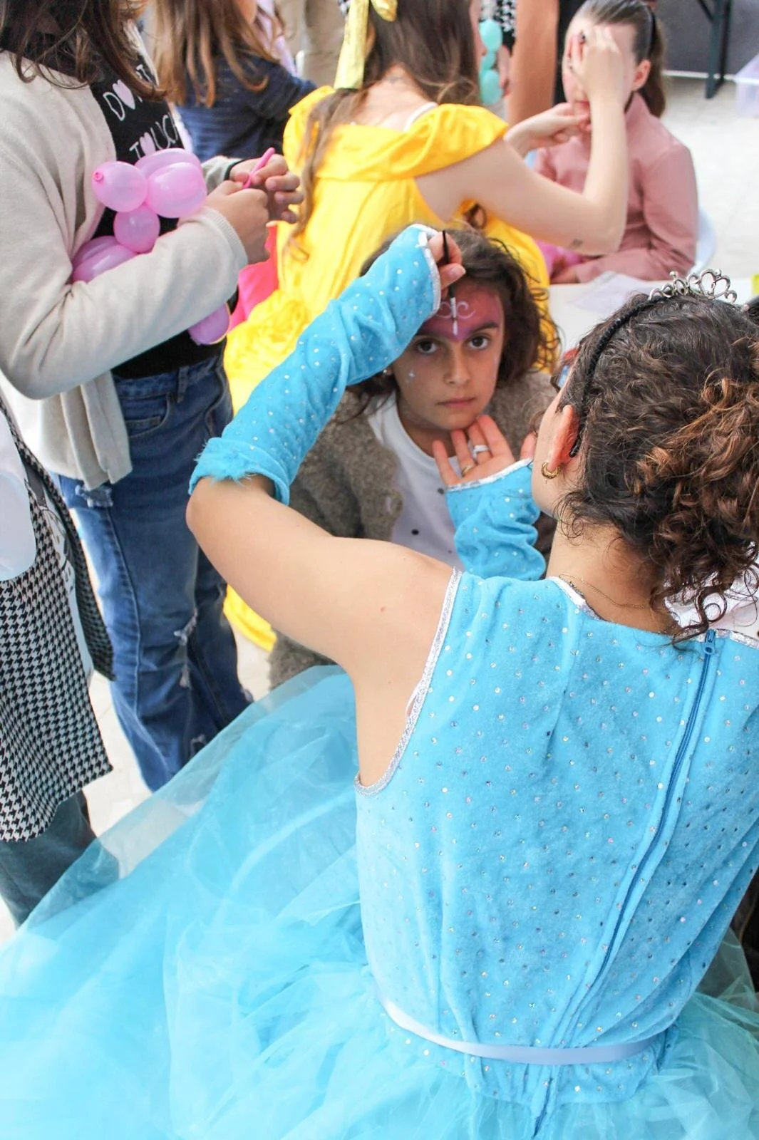 Danza di una ragazza in costume azzurro con davanti un trucca-faccia di una ragazza con i capelli ricci scuri e abbigliamento fantasia, in un evento di trucco per bambini.