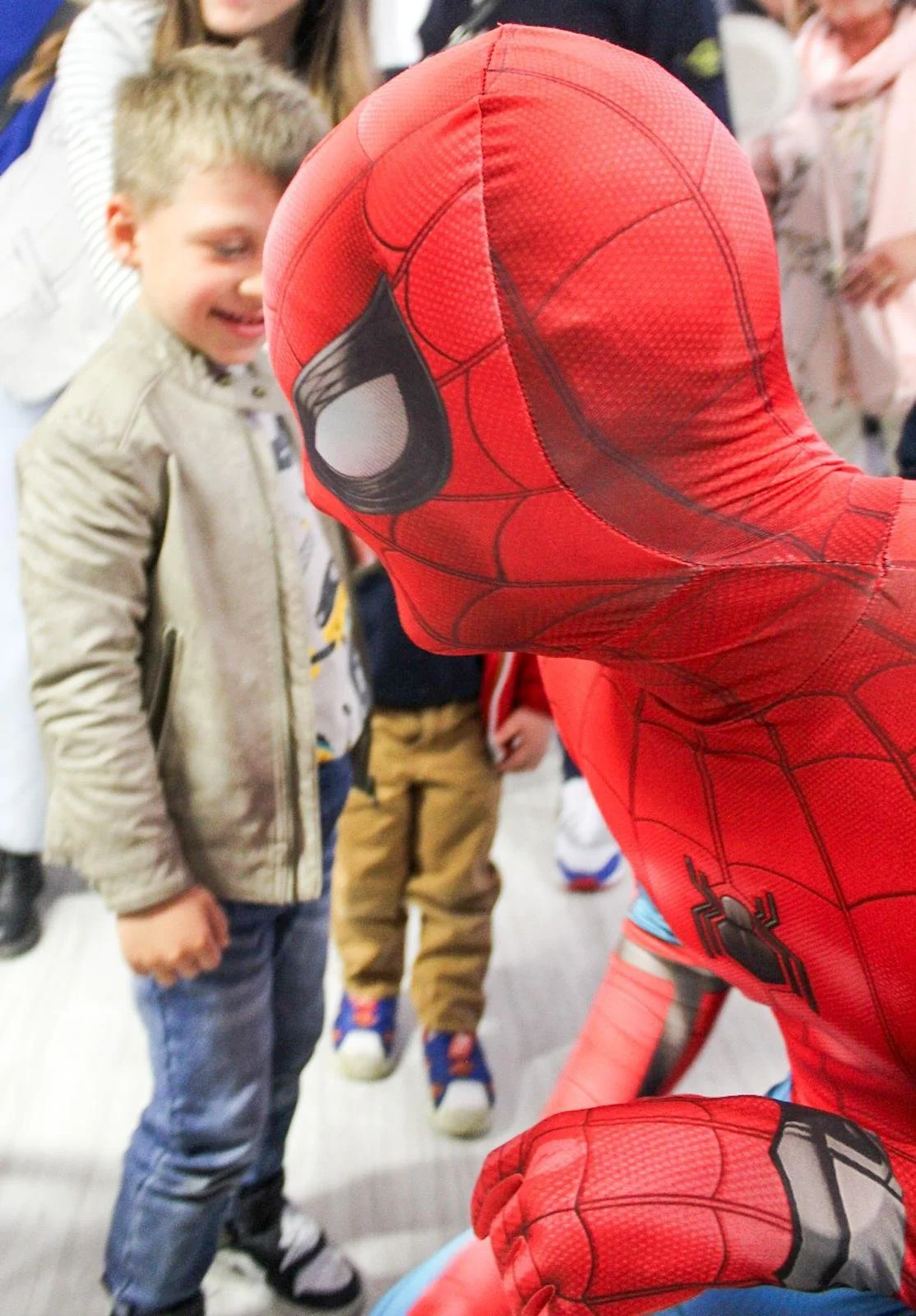 Ragazza vestita da Spider-Man mentre interagisce con un bambino sorridente, in un ambiente interno con altre persone sullo sfondo.