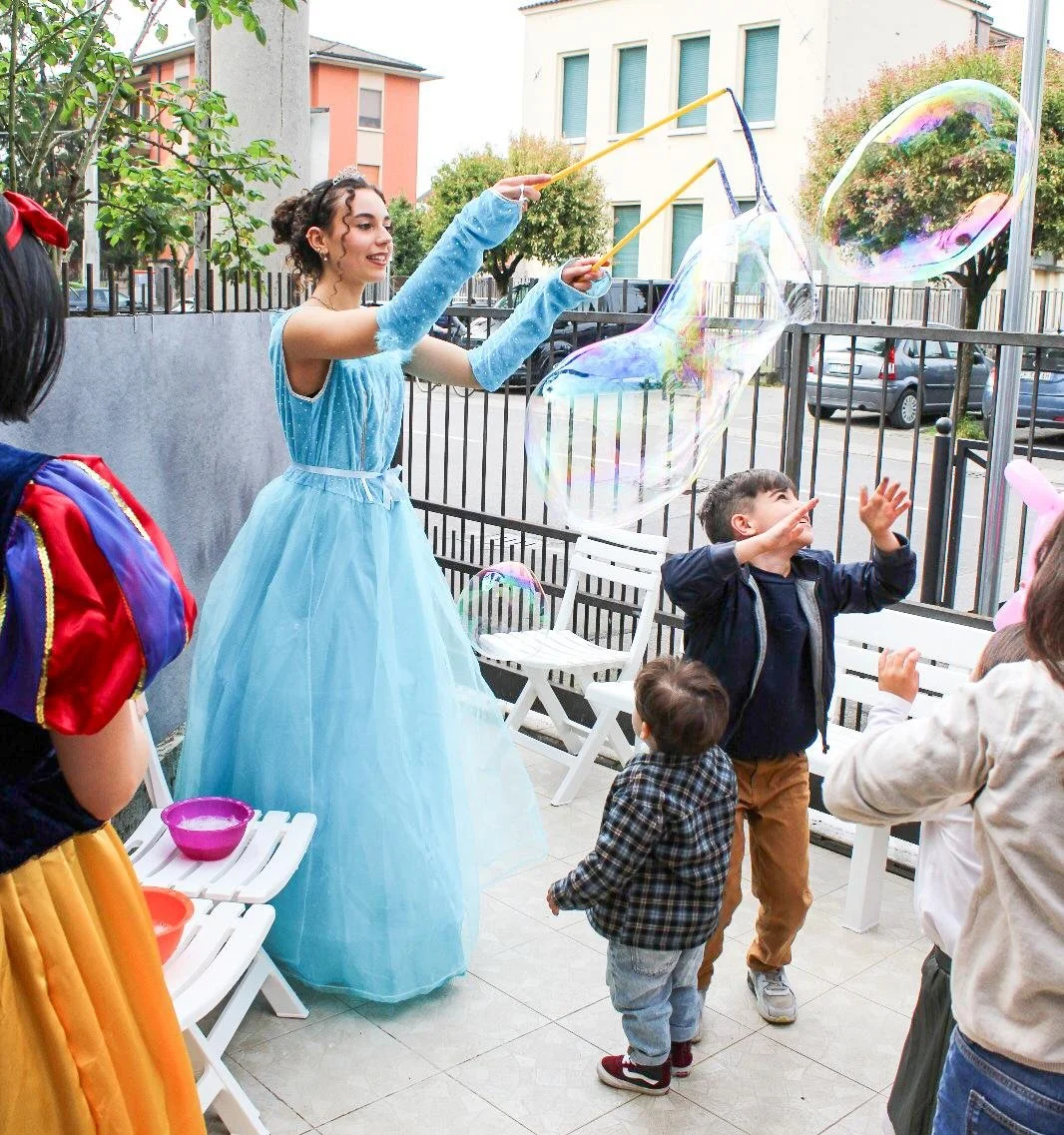 Una donna vestita da principessa azzurra fa fare bolle di sapone ai bambini in un cortile esterno con parete grigia e foresta di alberi.