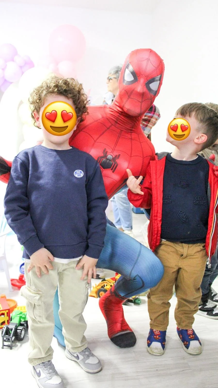 Due bambini sorridenti con emoticon faccia con occhi a cuore che si trovano accanto a una persona vestita da Spider-Man.