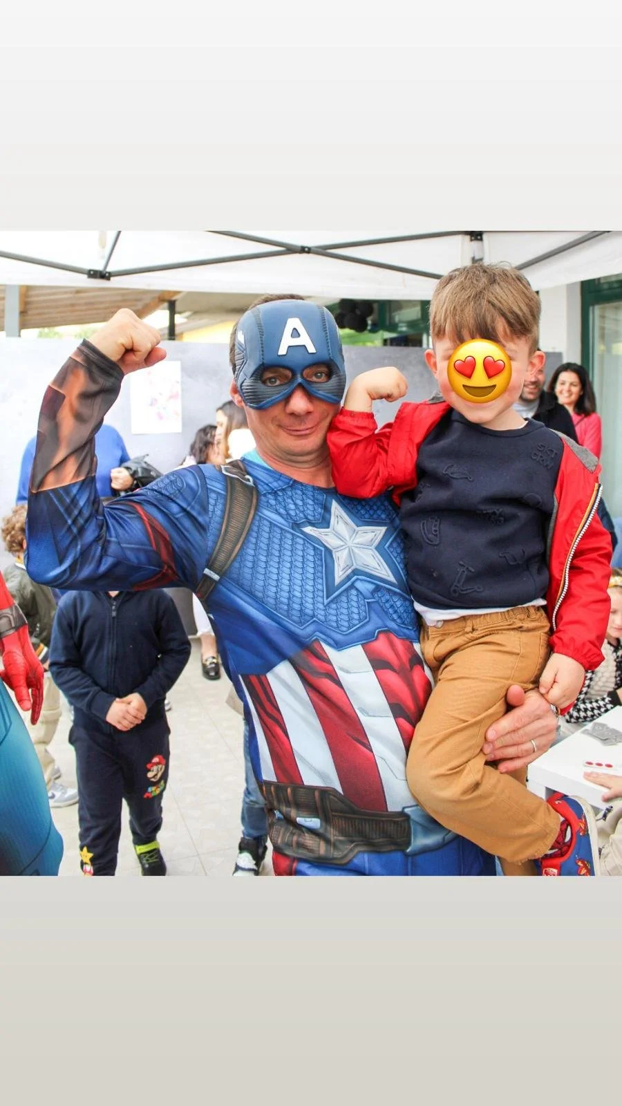 Un uomo vestito da Captain America tiene un bambino in braccio durante un evento pubblico, entrambi con espressione felice. Altri bambini e adulti sono visibili sullo sfondo.