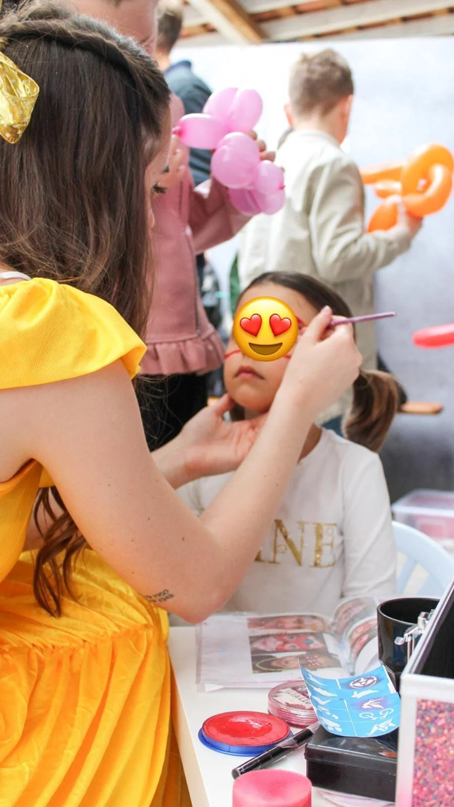 Una donna sta truccando una bambina con gli occhi coperti da un'emoji a cuore. Sul tavolo ci sono colori, pennarelli e album fotografici. In background, persone che fanno acconciature con palloncini.