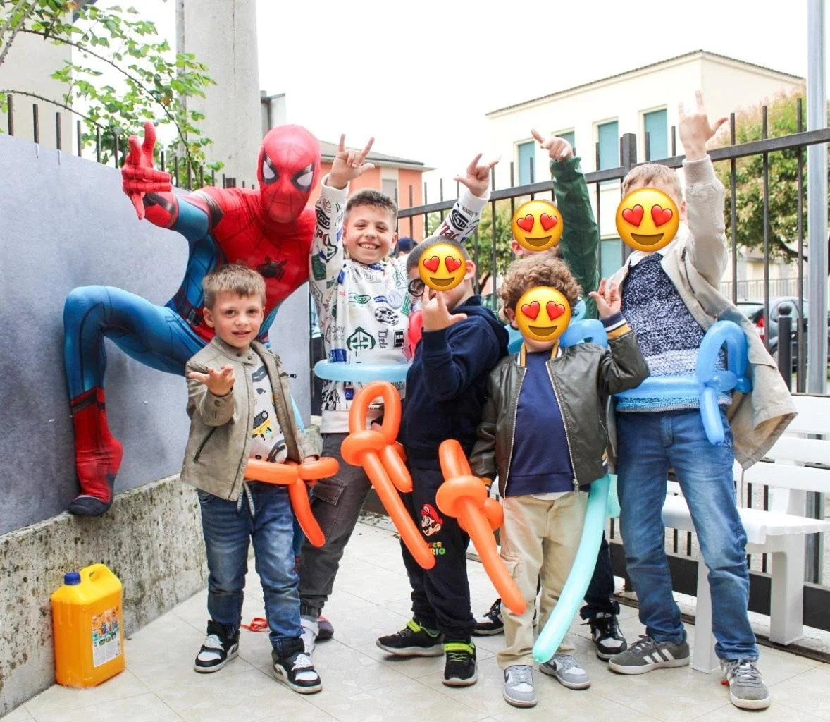 Gruppo di bambini felici con un uomo vestito da Spider-Man, alcuni fanno il gesto rock con le mani, alcuni indossano palloncini colorati, in un cortile esterno, con edifici e alberi sullo sfondo.