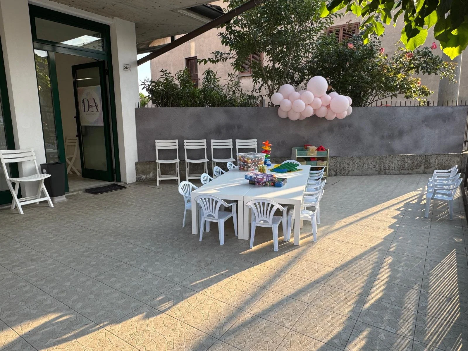 Spazio esterno all'aperto con tavolo bianco e sedie bianche, decorato con palloncini rosa e bianco, giochi per bambini, piante e edificio sullo sfondo.