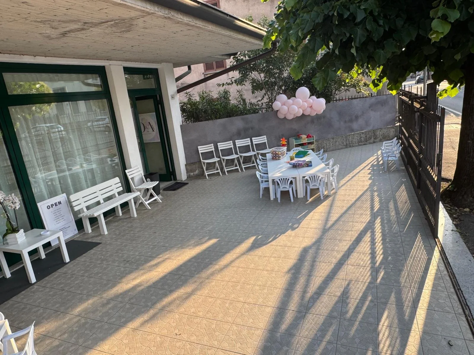 Veranda decorata per una festa, con tavolo e sedie bianche, palloncini rosa, decorazioni e un cartello 'OPEN'.