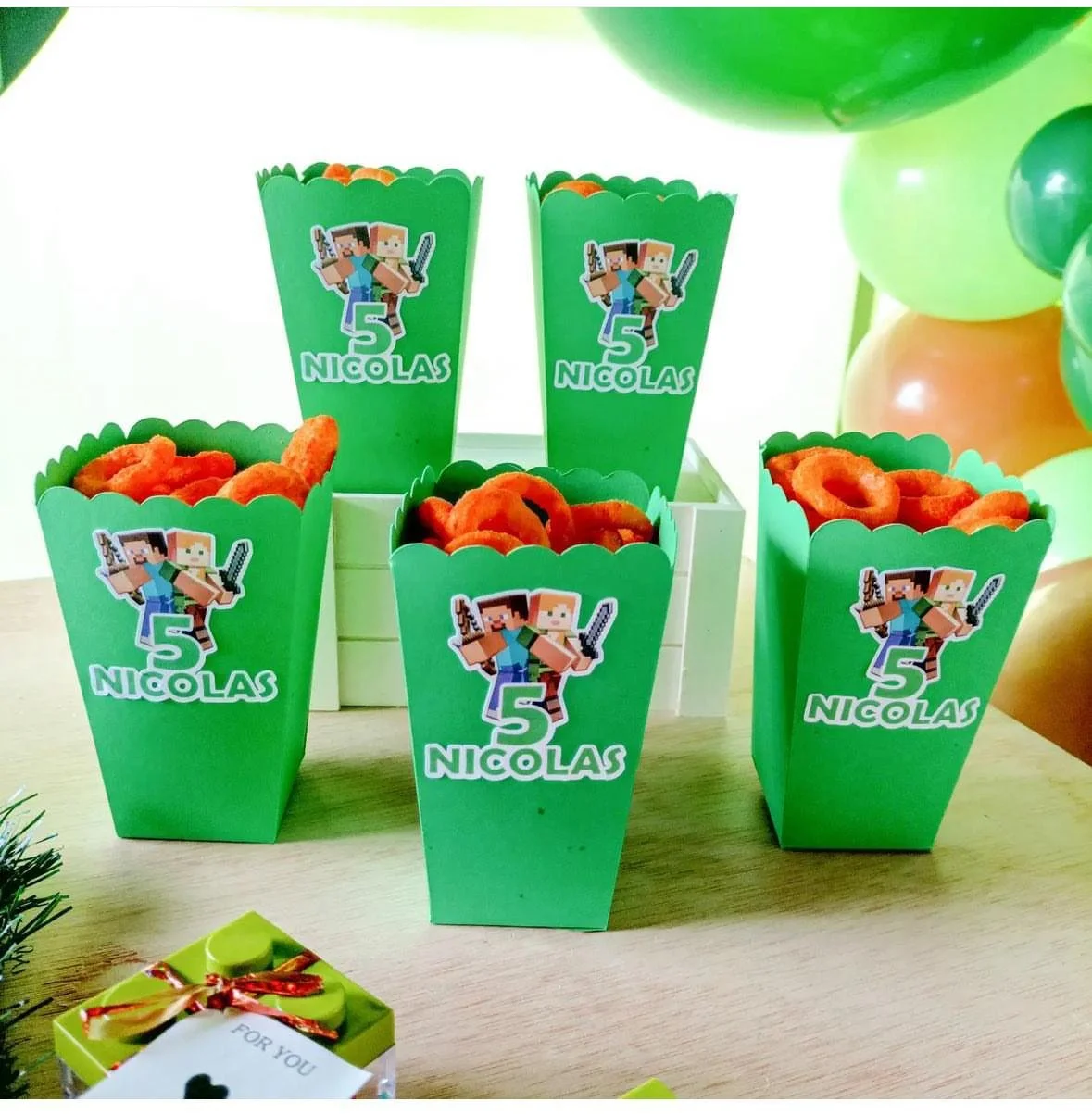 Contenitori verdi a forma di popcorn con etichette di Minecraft e il numero 5, pieni di snack arancioni, disposti su un tavolo decorato per una festa di compleanno.