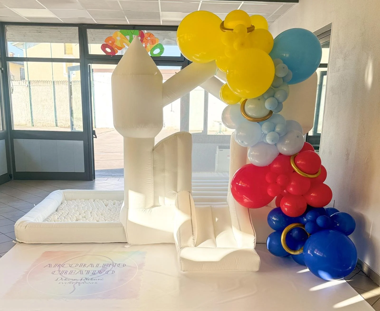 Decorazione con palloncini colorati e struttura in tessuto bianco, inclusa una grande lettera 'L', in una stanza con porte di vetro che danno all'esterno.