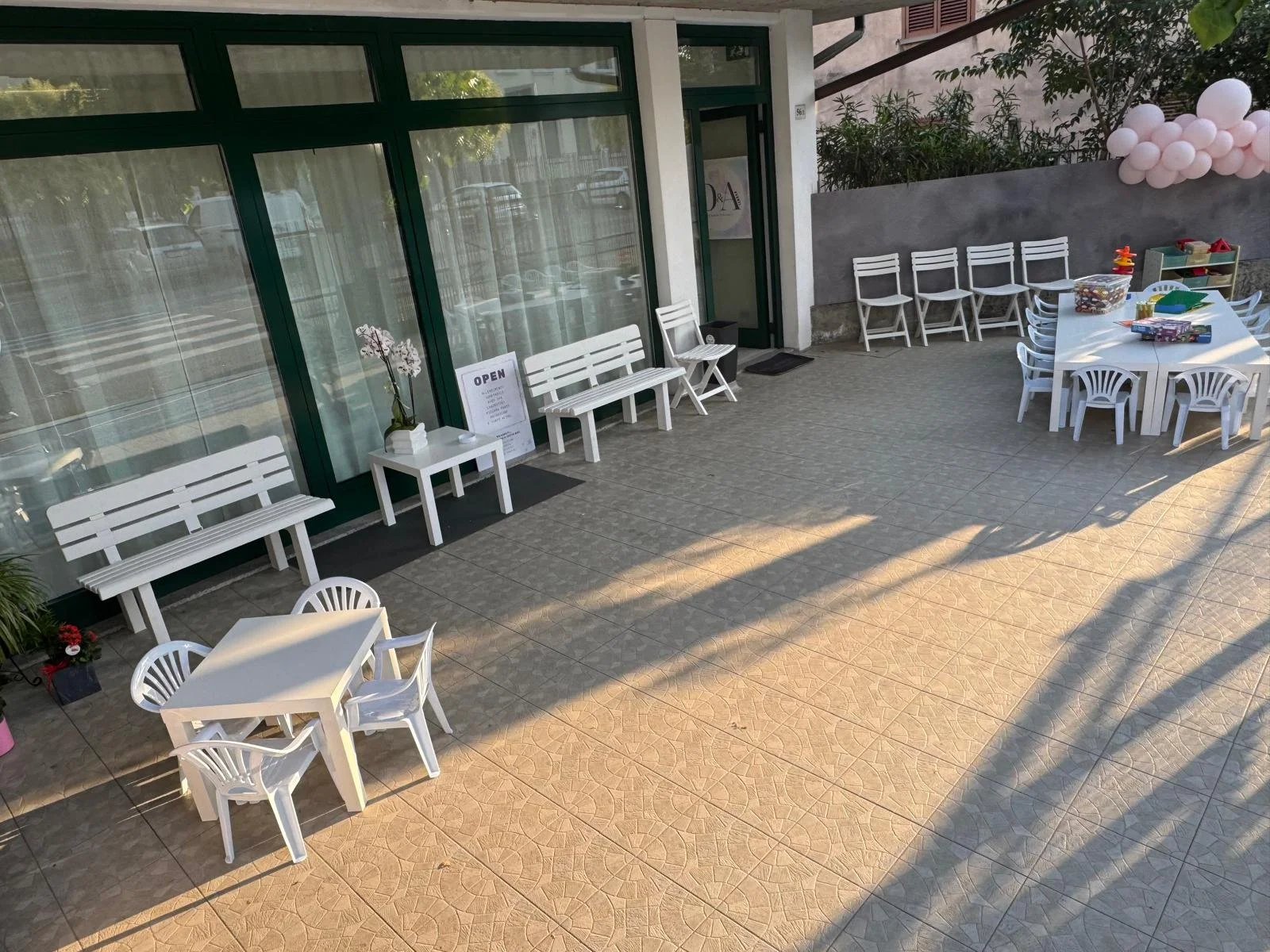 Spazio esterno di un locale con sedie e tavoli bianchi, qualche decorazione con palloncini rosa e piante, pareti di vetro e porta d'ingresso, in ombra con abbondante luce solare.