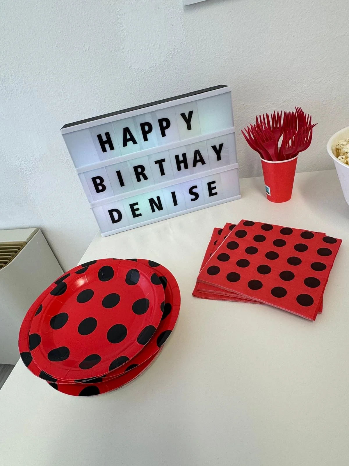 Decorazioni per una festa di compleanno a tema ladybug con piatti rossi e neri a pois, tovaglioli, forchette, e una scatola luminosa con scritto "Happy Birthday Denise".