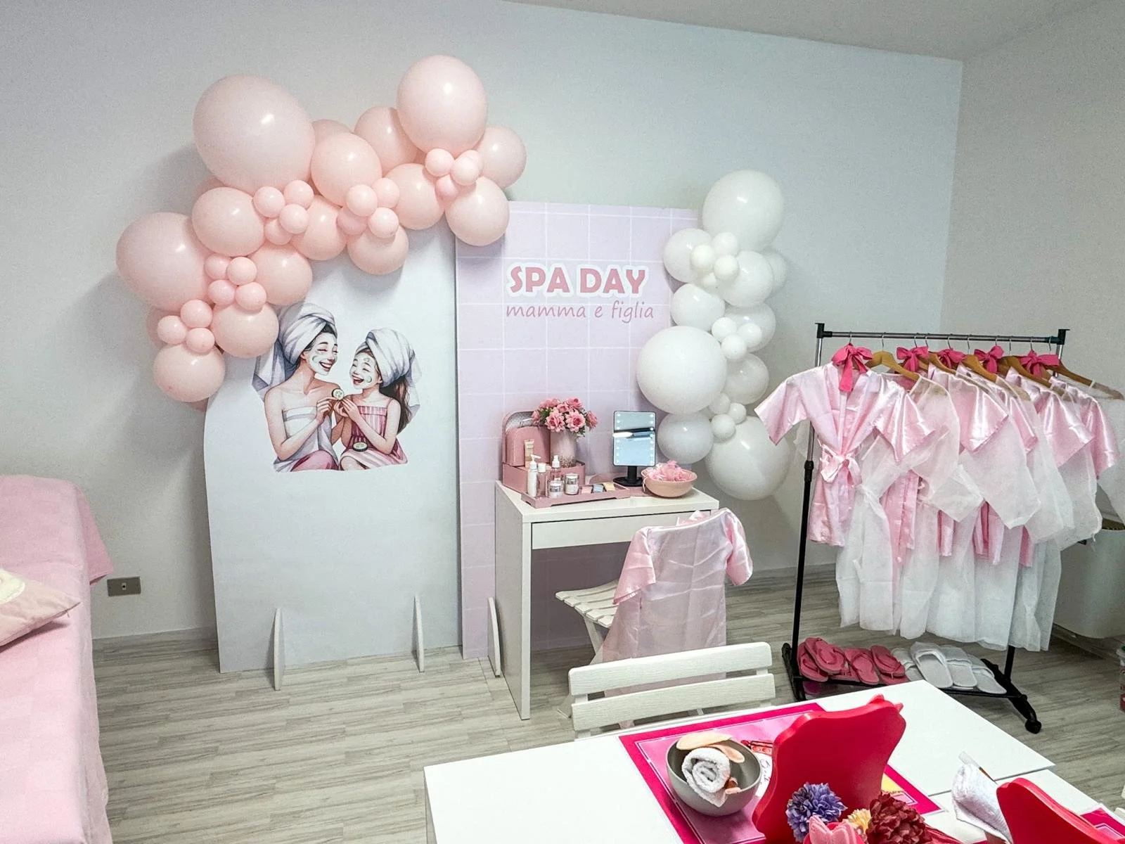 Decorazione per la giornata della mamma e figlia con palloncini rosa e bianchi, banner con scritto SPA DAY mamma e figlia, vestiti rosa e bianchi appesi, tavolo con fiori e prodotti di bellezza