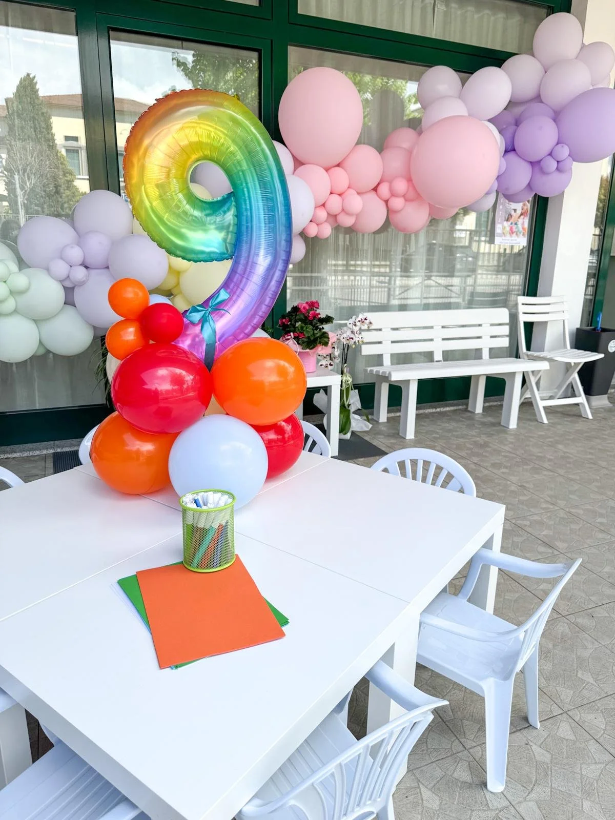 Decorazione di festa con palloncini colorati, tra cui un grande palloncino a forma di numero 9 con arco iris, e tavolo con fogli colorati e contenitore di penne.