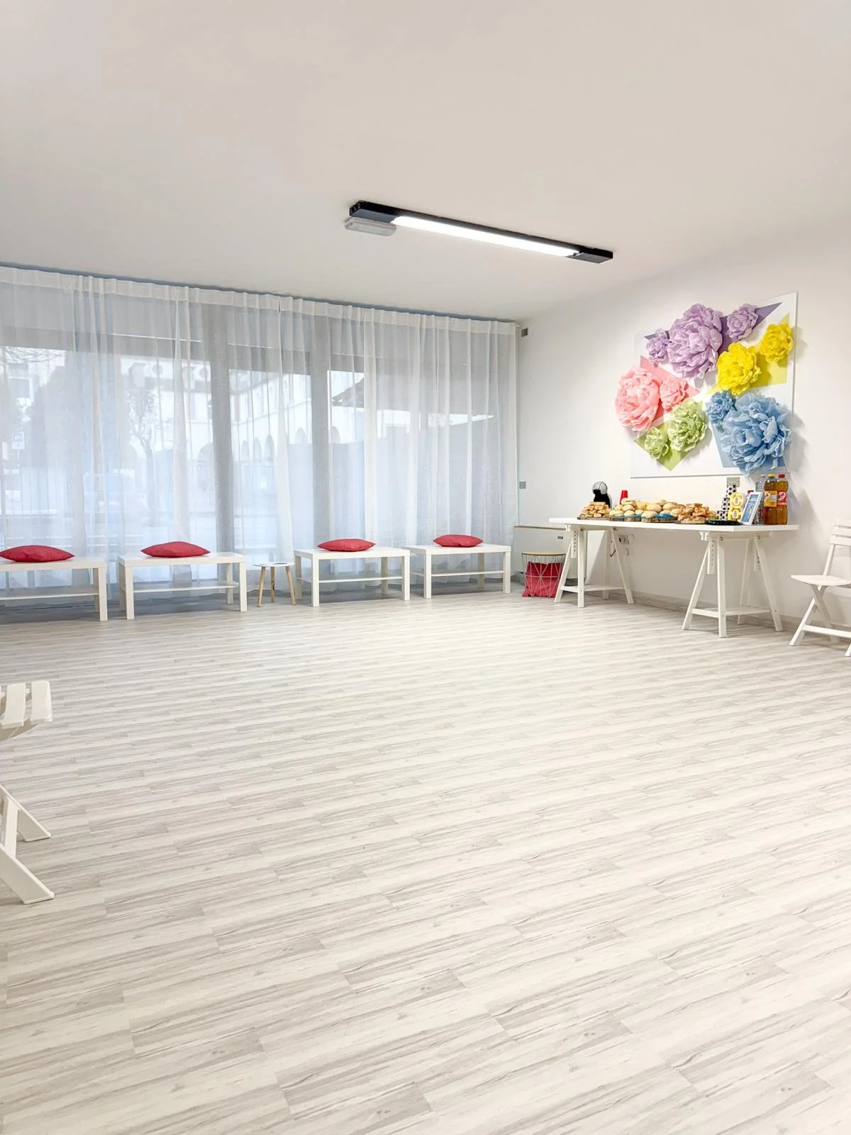 Una stanza vuota con pareti bianche, pavimento chiaro, tende trasparenti alle finestre, una decorazione floreale colorata sulla parete, e un tavolo con snack e bevande.