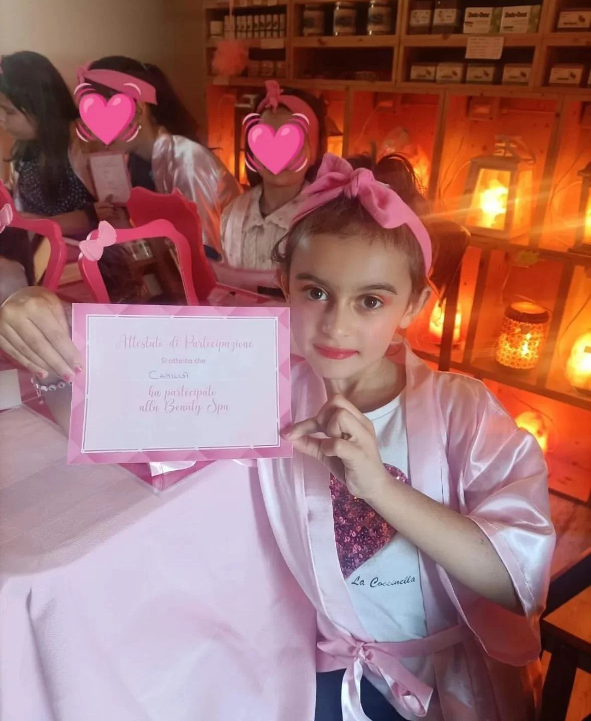 Una bambina che tiene un certificato di partecipazione alla Beauty Spa, indossa una maglietta bianca con un cuoricino e una vestaglia rosa, con un fiocco rosa in testa, seduta a un tavolo in un locale decorato con luci calde.