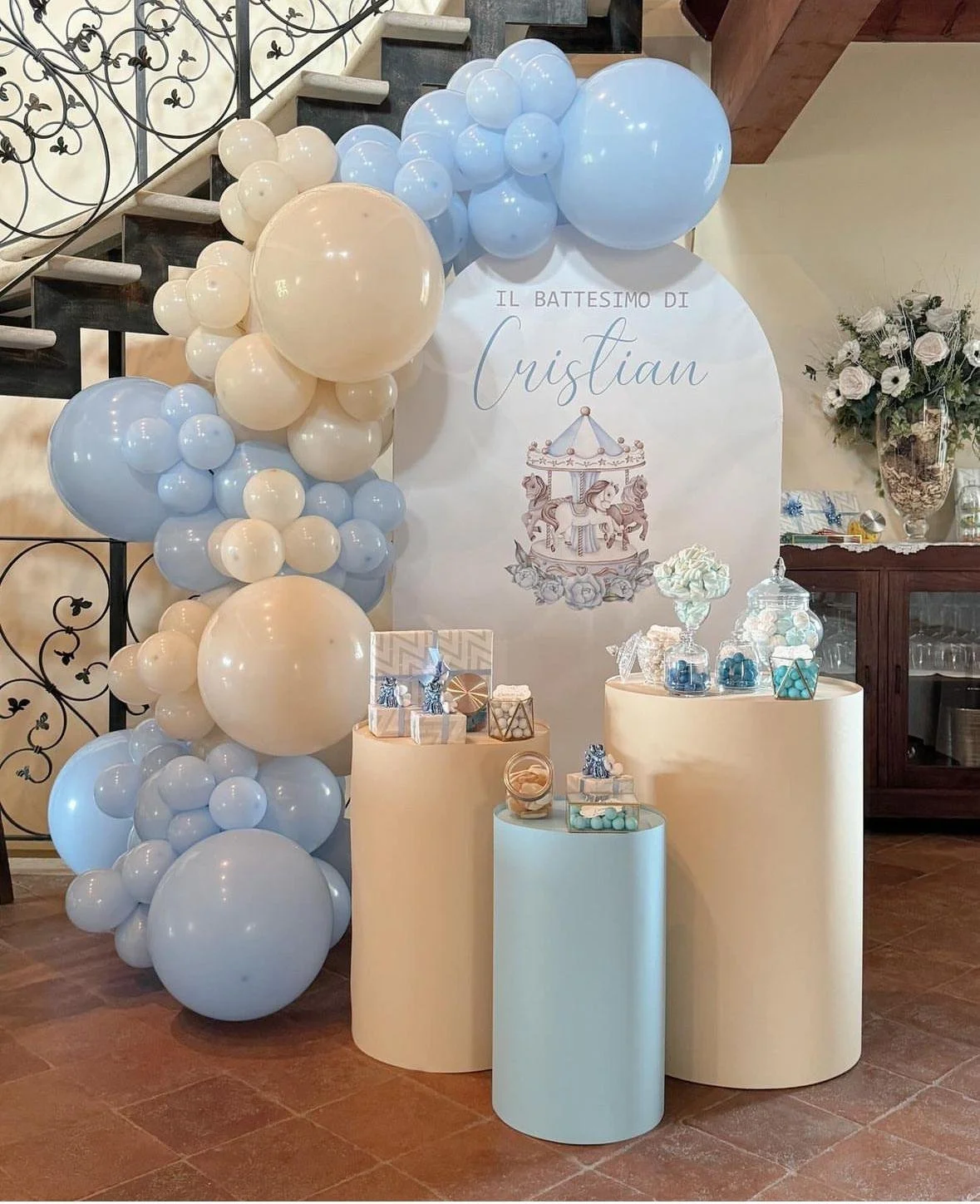 Decorazioni per il battesimo di Cristian, con palloncini nei colori bianco e azzurro, un banner con il testo in italiano, e una mostra di dolci e regali.