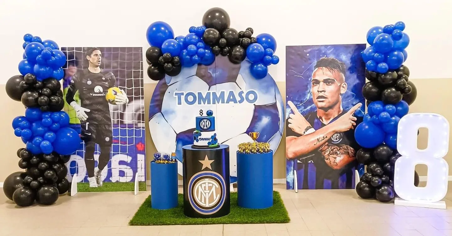 Decorazioni e fotografie in stile calcio per il compleanno di Tommaso, con palloncini blu e neri, una torta a due livelli con temi calcistici, e gigantografie di due calciatori.
