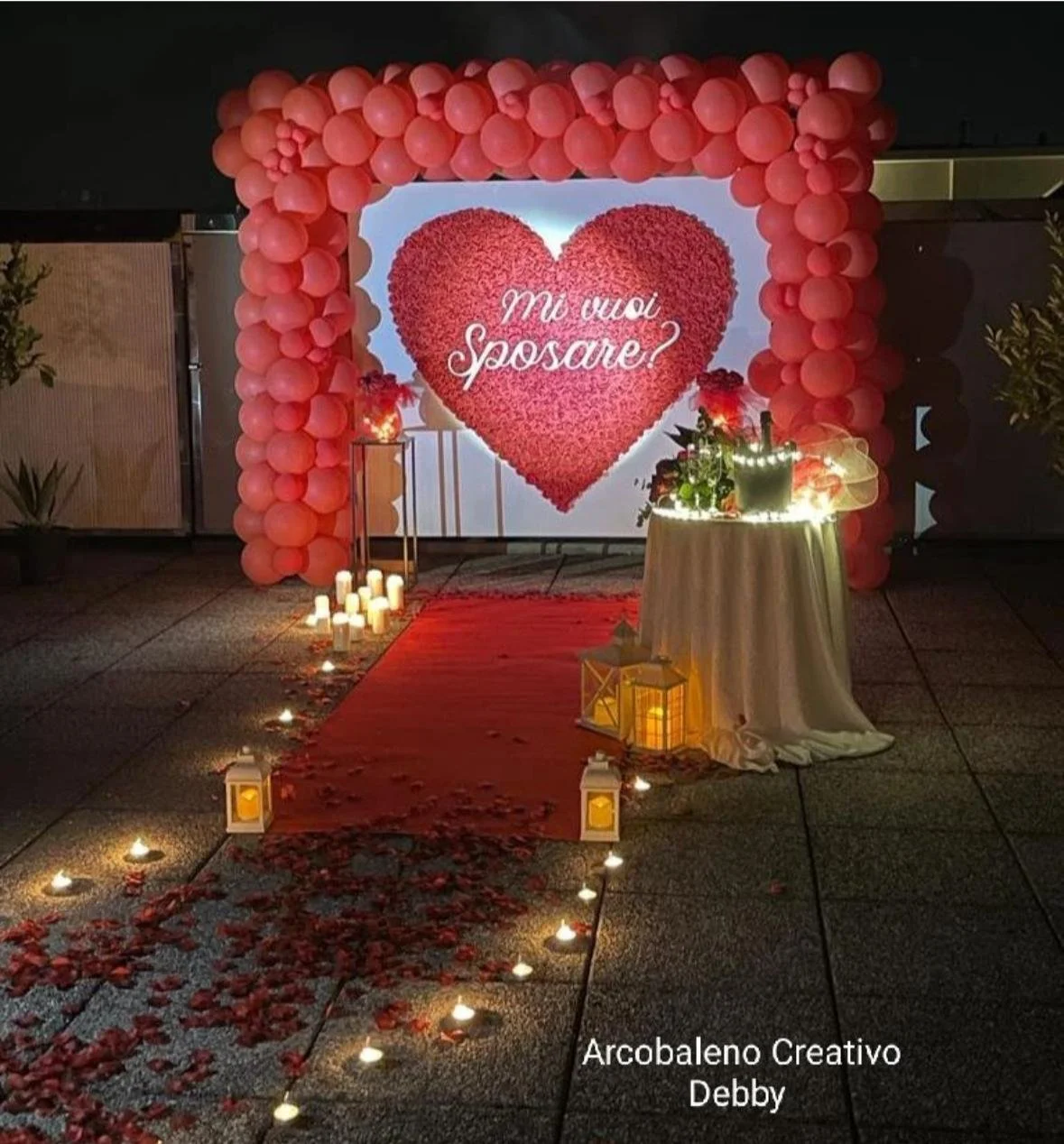 Decorazione per proposta di matrimonio con grande cuore di fiori rossi e sfondo con palloncini rosa, sentiero di petali e candele. La scritta sullo sfondo dice "Mi vuoi sposare?".