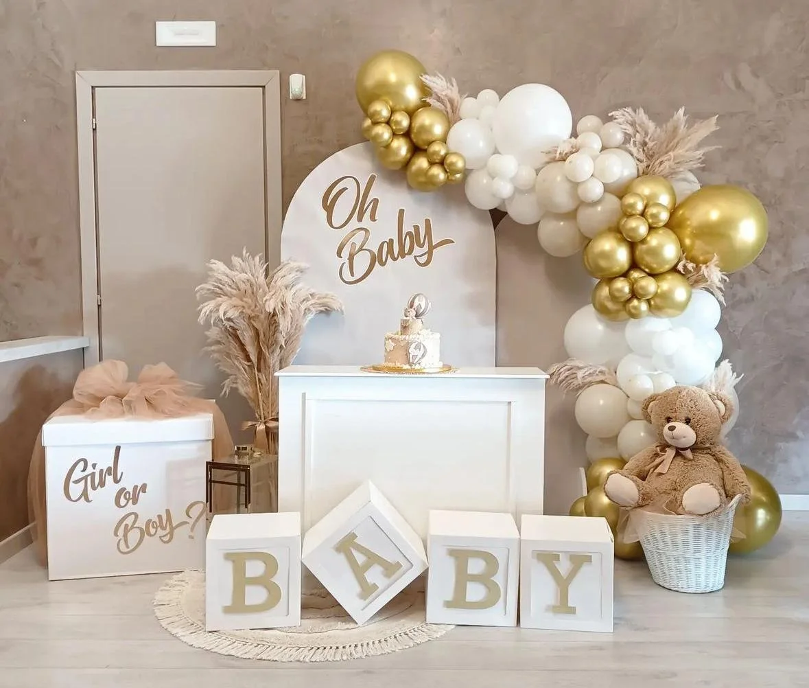 Decorazione per festa di nascita con palloncini oro e bianchi, teddy bear, blocchi con lettere B A B Y, e cartello con scritto 'Oh Baby'.