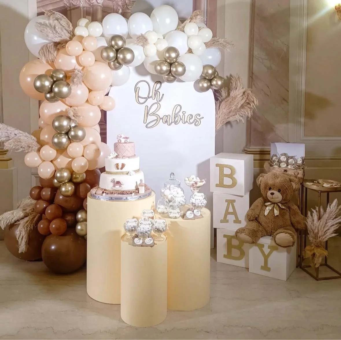 Decorazione per baby shower con palloncini rosa, oro e bianco, torta, teddy bear e decorazioni sabaidee con le lettere BABY. Sfondo con scritta 'Oh Babies'.