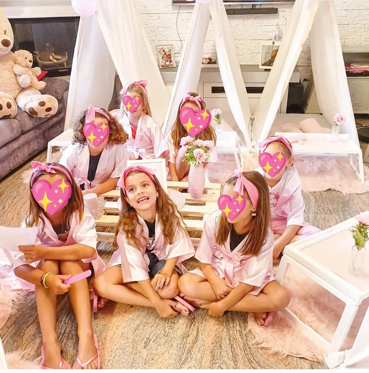 Gruppo di bambine che partecipano a una festa, vestite con camicette rosa e accessori, sedute su un tappeto e intorno a tavolini decorati con fiori