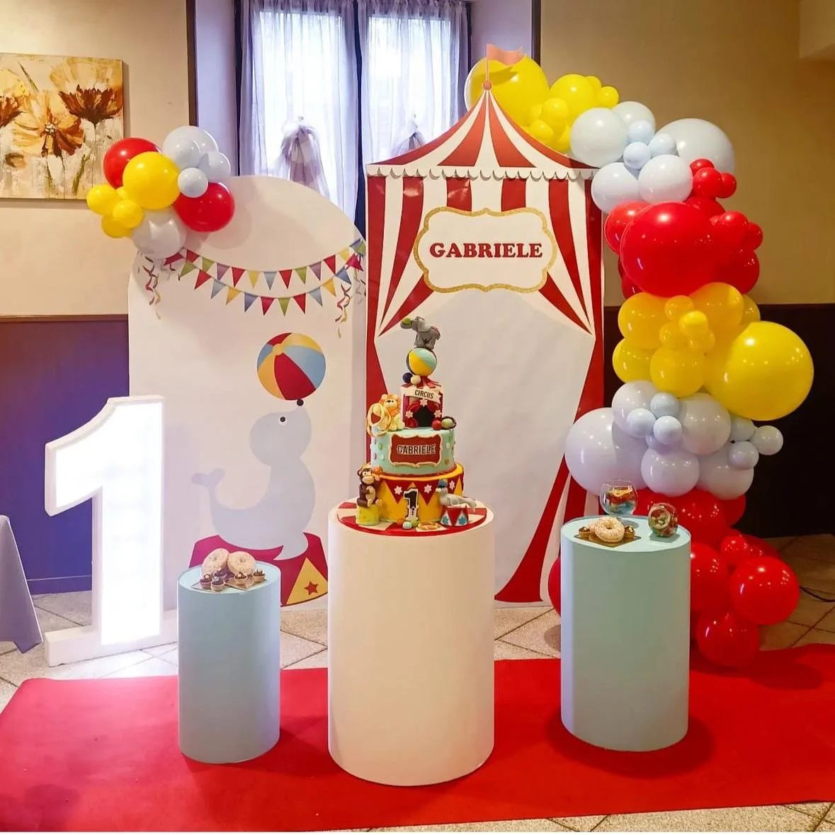 Decorazioni per festa di compleanno con tema circus, including palloncini rossi, gialli e bianchi, un grande numero 1, una torta decorata e uno sfondo a strisce rosse e bianche con il nome Gabriele.